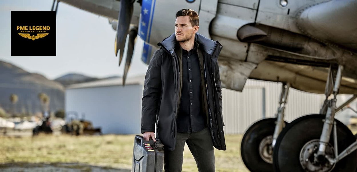 Parka noire avec capuche bordée de fourrure, portée par-dessus une chemise sombre à boutons, debout à côté d'un avion, tenant une mallette à outils rectangulaire.