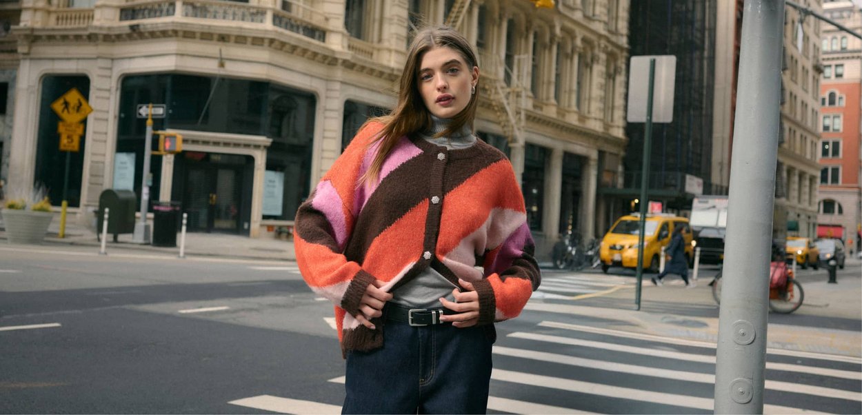 Flerfärgad stickad kofta med orange, rosa, brun och vit geometriska mönster. Knäppt fram och lösa ärmar, kombinerad med en polotröja.