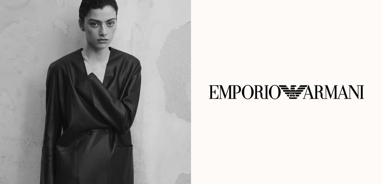 Mujer con blazer de cuero negro de pie contra una pared desgastada, junto al logo de Emporio Armani sobre un fondo blanco liso.