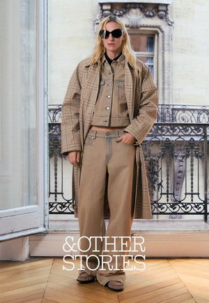 Lyshåret kvinde iført storternet frakke, tan denimjakke og bukser med vide ben, står indendørs nær udsmykket balkongelænder.