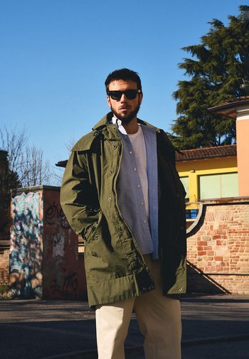 Olijfgroene parka jas met een gestructureerde afwerking, gedragen over een lichtblauw shirt. Het model draagt donkere zonnebrillen en crèmekleurige broek.