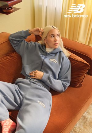 Lichtblauwe hoodie met rits en geborduurd logo, gecombineerd met bijpassende joggingbroek. Zachte stoftextuur, ontspannen pasvorm, liggend op een bruine bank.