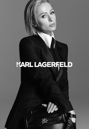 Chaqueta negra con textura satinada, cuello blanco, sosteniendo un bolso de cuero negro con herrajes plateados. El texto debajo dice "KARL LAGERFELD."