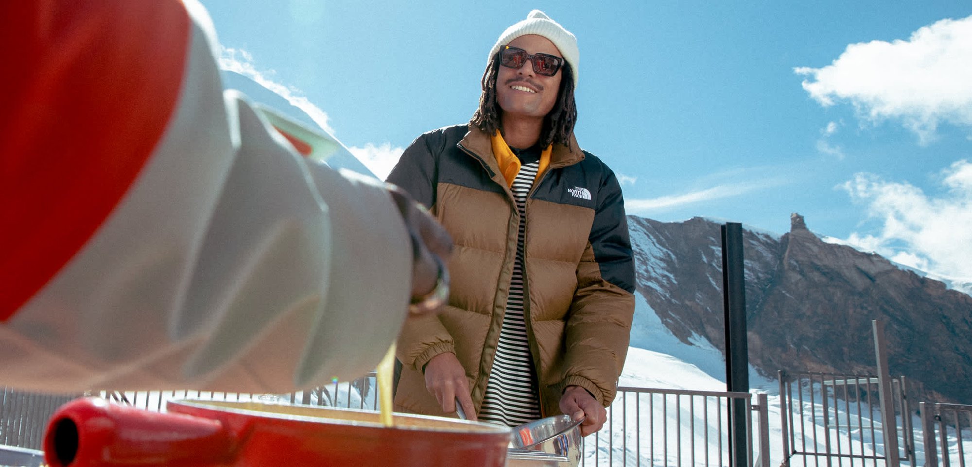 Person i vinterjakke og hue, der rører mad udendørs med bjerge og en klar sol i den klare himmel bag dem.
