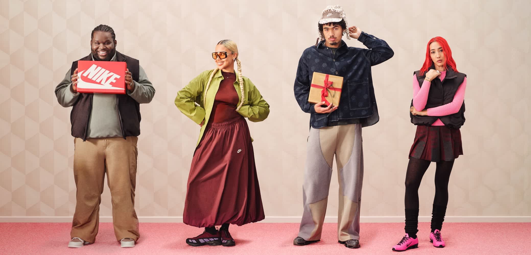 Quattro persone sono in piedi davanti a uno sfondo chiaro. Un uomo tiene una scatola rossa di scarpe Nike, una donna indossa una giacca verde e una gonna color bordeaux, un altro uomo con una camicia a motivi tiene un regalo incartato e una donna indossa un gilet nero sopra una maglietta e una gonna rosa.