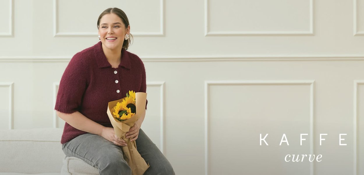 Burgunderfarbener Pullover mit kurzen Ärmeln, Kragen und Knopfdetails. Das Modell hält einen Strauß gelber Sonnenblumen, der in Kraftpapier eingewickelt ist.