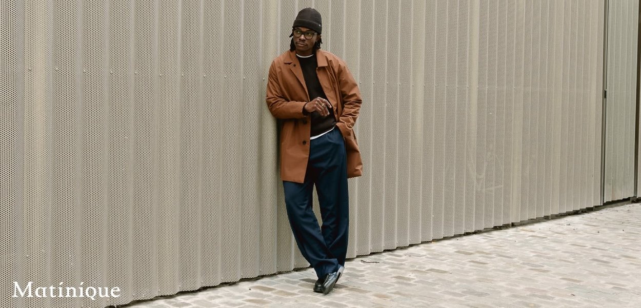 Brauner Mantel über einem schwarzen Pullover, marineblaue Hose und schwarze Schuhe. Das Modell lehnt an einer strukturierten, perforierten Wand.