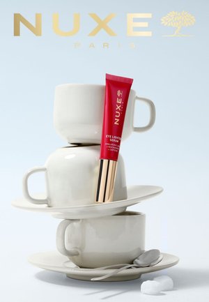 Tubo del siero illuminante per occhi rosso NUXE appoggiato su tazze e piattini bianchi impilati, con un cucchiaio e due zollette di zucchero accanto.
