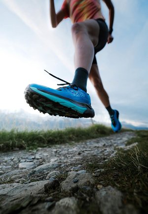Bottine de trail de course bleue et noire avec une semelle texturée, des lacets noués et une marque visible, positionnée sur un chemin en gravier.