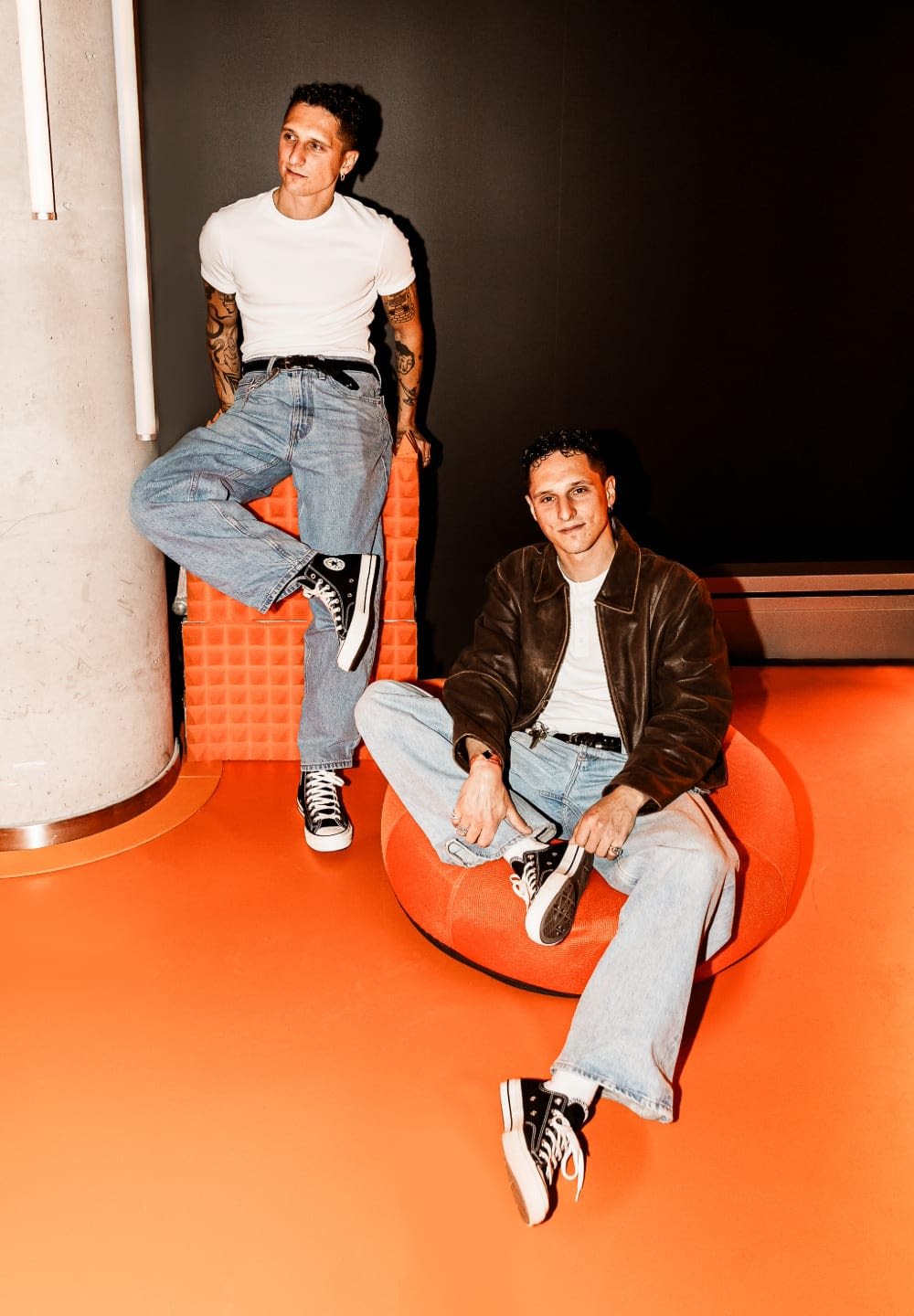 Two individuals in casual outfits. One wears a white t-shirt and blue jeans; the other a brown jacket over a white top. Both have black sneakers.