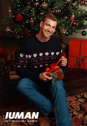 Pull en tricot avec des motifs festifs de Père Noël, associé à un jean bleu. Tenant un petit cadeau vert et rouge emballé avec un ruban près d'un sapin de Noël décoré.