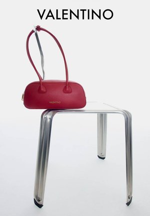 Sac à main Red Valentino avec doubles poignées reposant sur un tabouret en métal minimaliste sur fond blanc.