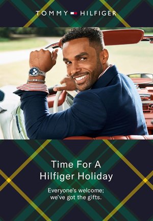 Werbebild von Tommy Hilfiger mit einem lächelnden Mann, der einen marineblauen Blazer mit gestreiften Manschetten und eine blaue Uhr trägt und in einem Oldtimer sitzt.