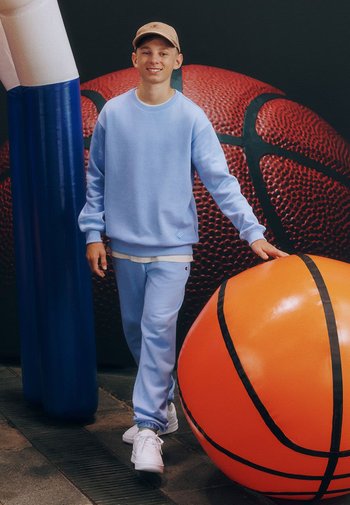 Helles blaues Sweatshirt und passende Jogginghose, weiße Sneakers, stehend neben einem übergroßen orangefarbenen Basketball und aufblasbarem Sportgerät.