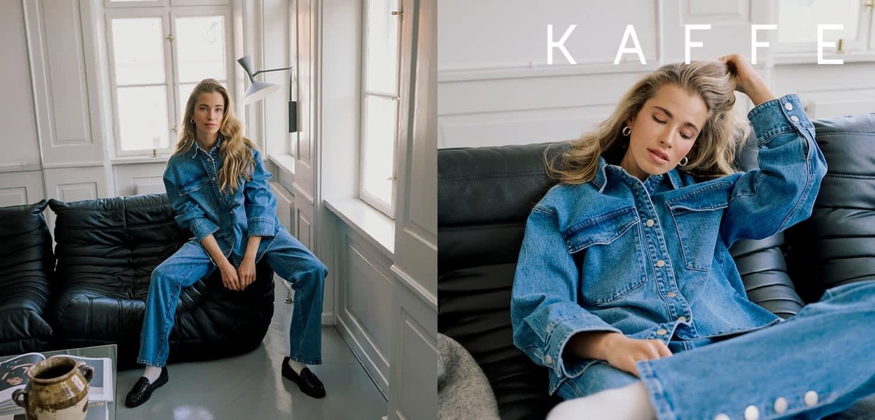 Junge Frau in blauem Jeansjacke und Jeans posiert und entspannt sich auf einem schwarzen Ledersofa in einem hellen, weißwandigen Raum mit großen Fenstern.