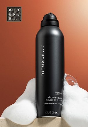 Conteneur cylindrique noir étiqueté "RITUALS" avec une buse, placé sur une surface crème texturée avec de la mousse blanche et des bulles, sur fond rouille.