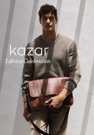 Brown leather messenger bag with a flap, adjustable strap, and metal hardware. Features embossed logo. Model wearing a light sweater and pants.