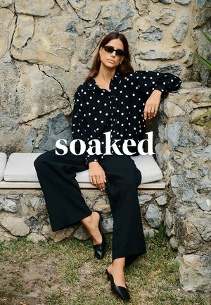 Schwarze Bluse mit Polka-Dots und lockeren Ärmeln, kombiniert mit weiten schwarzen Hosen und eleganten schwarzen Mules, vor einer strukturierten Steinwand.