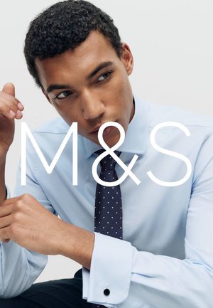 Man wearing light blue dress shirt and navy blue polka dot tie, resting arm on knee against plain background.