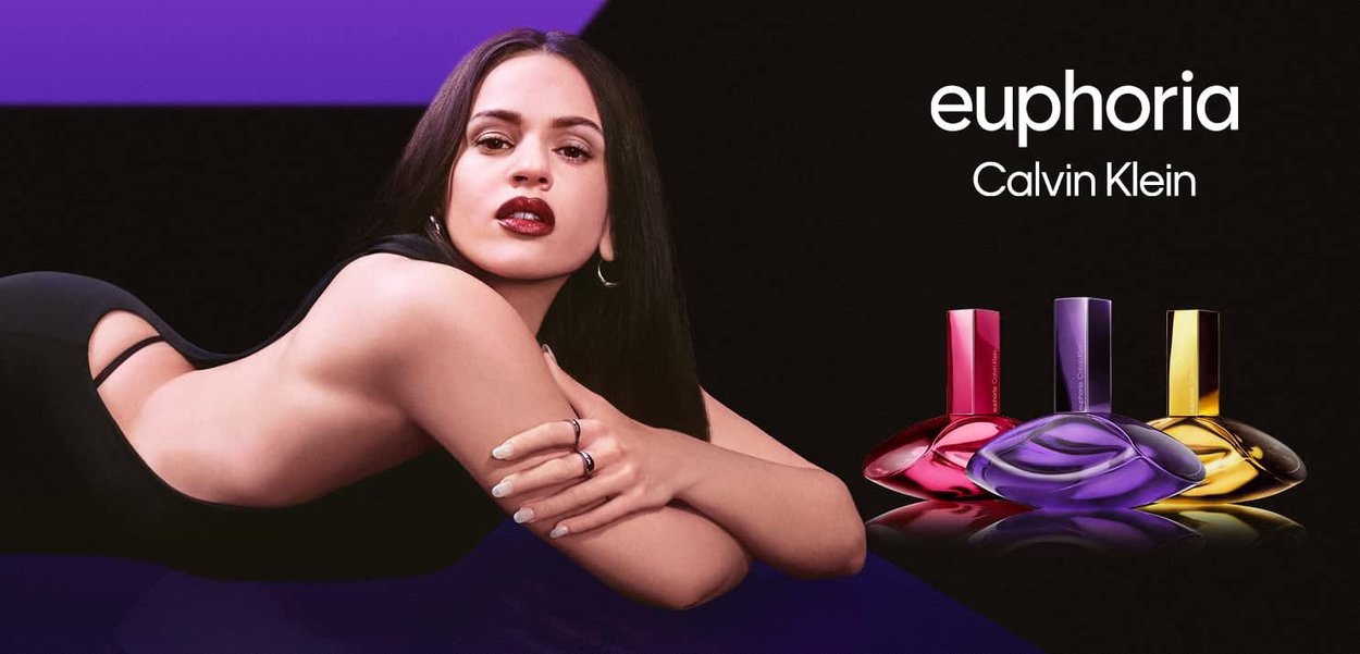Woman with long dark hair and dark lipstick reclining next to three colorful Calvin Klein Euphoria perfume bottles on a black background.