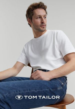 Weißes Baumwoll-T-Shirt mit kurzen Ärmeln, kombiniert mit dunkelblauen Jeans. Das Model sitzt auf einem Holzstuhl.