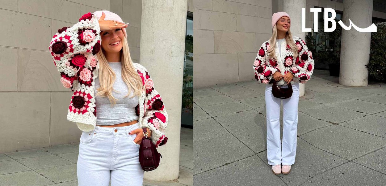 Vrouw in een roze beanie, een gebloemde gebreide cardigan, een grijze crop top, een witte broek en roze schoenen staat op de stoep bij betonnen kolommen.
