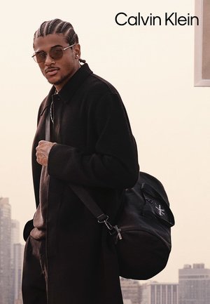 Cappotto nero sopra un maglione scuro, indossando occhiali da sole rotondi. Una zaino nero con un logo poggia sulla sua spalla. Skyline urbano sullo sfondo.