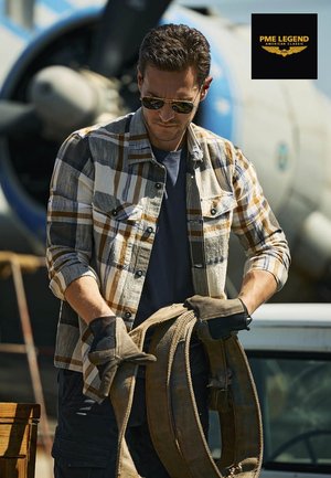 Ein Mann mit Sonnenbrille und Handschuhen hantiert mit dicken Gurten vor einem Oldtimer-Flugzeug mit Propeller im Hintergrund.