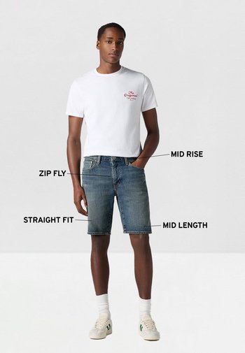 Camiseta blanca con texto rojo combinada con pantalones cortos de mezclilla azul, de tiro medio, corte recto, con cremallera y de longitud media, llevados con calcetines blancos y zapatillas.