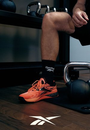 Persoon draagt oranje Reebok-schoenen en zwarte sokken, zittend bij een kettlebell en een rek met kettlebells op een houten vloer.