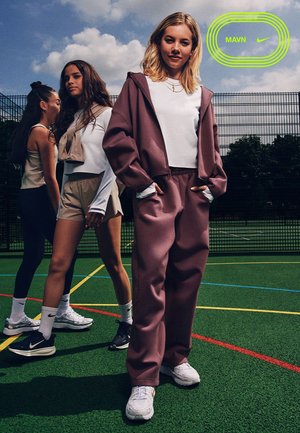 Drei junge Frauen in lässiger Sportkleidung auf einem Außenplatz, eine in einem mauvefarbenen Trainingsanzug mit Händen in den Taschen, die anderen in Shorts und Turnschuhen.