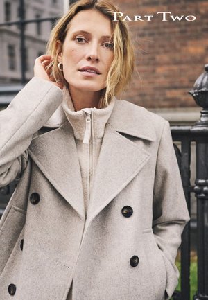 Manteau beige à double boutonnage avec un large col, agrémenté de boutons foncés et d'une couche intérieure zippée à col montant. Texture de tissu douce.