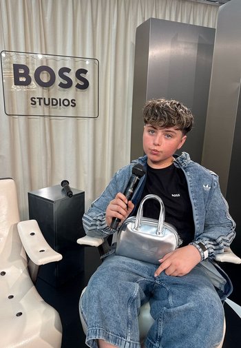 Jeune personne en tenue en jean tenant un micro et un sac argenté, assise dans un studio avec le logo BOSS Studios et une chaise blanche vide à proximité.