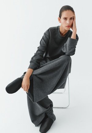 Grauer Strickpullover und weit geschnittene Hosen mit gerafften Bündchen, kombiniert mit schwarzen Schuhen. Modell sitzt auf einem transparenten Stuhl vor weißem Hintergrund.