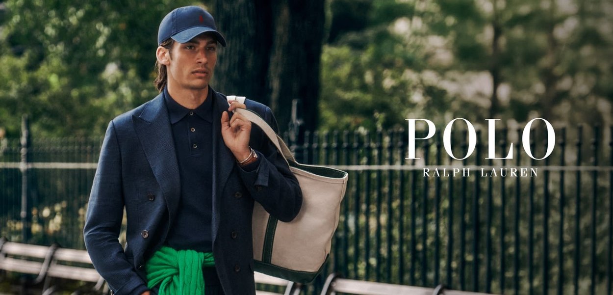 Mann i marineblå blazer og caps bærer beige totebag over skulderen med grønn genser knyttet rundt livet i en park.