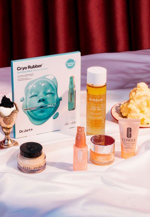 Masque apaisant Cryo Rubber dans un emballage bleu sarcelle, accompagné de divers produits de soin de la peau dans des contenants transparents ou pastel, sur un fond en tissu blanc.