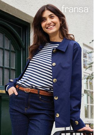 Frau lächelt im Freien, trägt eine marineblaue Jacke über einem blau-weiß gestreiften Hemd mit dunklen Jeans und einem braunen geflochtenen Gürtel.