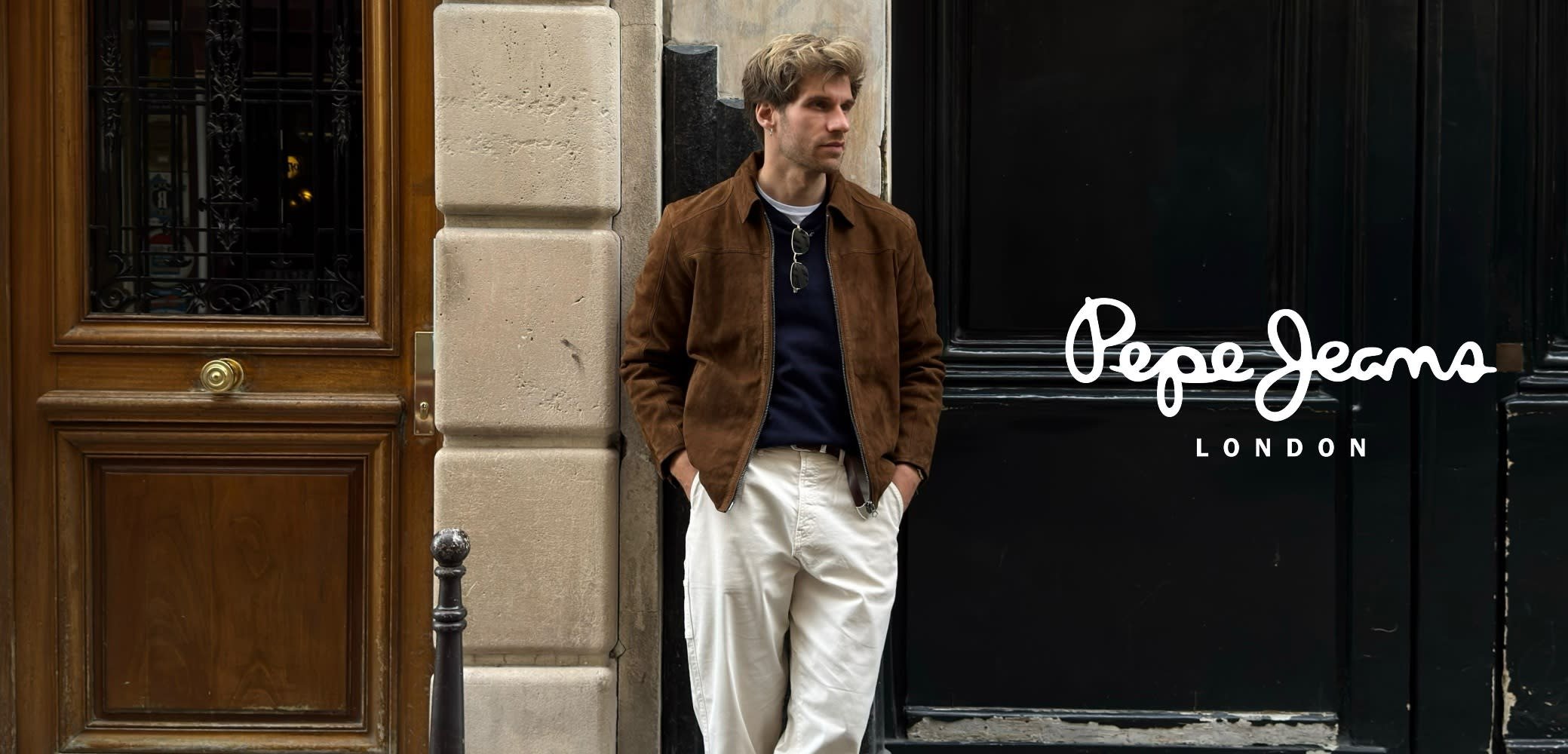 Bruine suède jas over een marineblauwe trui, witte broek en zwarte zonnebril, leunend tegen een muur met houten deur en metalen beslag.