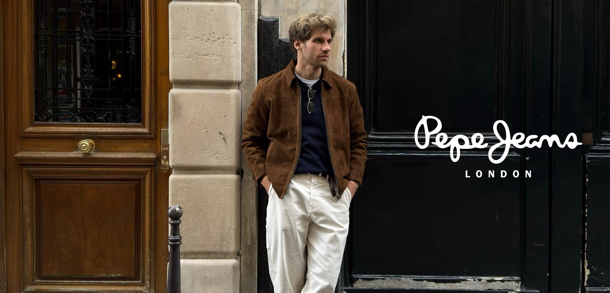 Giacca in camoscio marrone, maglione blu navy, pantaloni crema e stivaletti marrone scuro. In piedi contro un muro di pietra con una porta di legno.