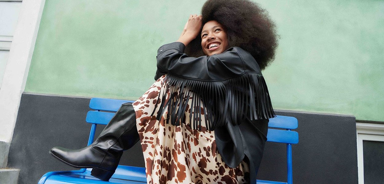 Black leather fringed jacket, beige dress with brown animal print, black knee-high boots, seated on a blue bench against a green background.