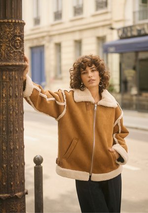 Brun jacka med foder av fårskinn, dragkedja, kontrasterande krämfärgad kant och sidofickor. Modellen lutar sig mot en dekorativ pelare.