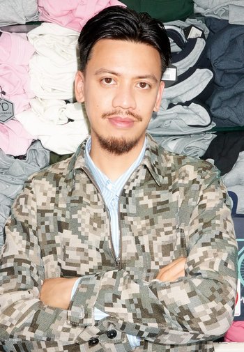 Camouflage-jack in groen- en grijstinten, met een ritssluiting. Lichtblauw shirt eronder. Achtergrond met opgevouwen kledingstapels.