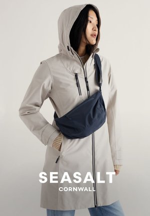 Kvinde iført beige hættejakke og marineblå crossbody-taske, stående med den ene hånd i lommen og kigger til siden mod en ensfarvet baggrund.