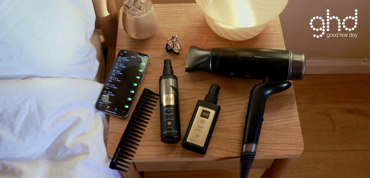 Nachtkastje met haardroger, kam, twee ghd haarverzorgingsflessen, smartphone met alarmen, kaars en schelp op houten vloer.