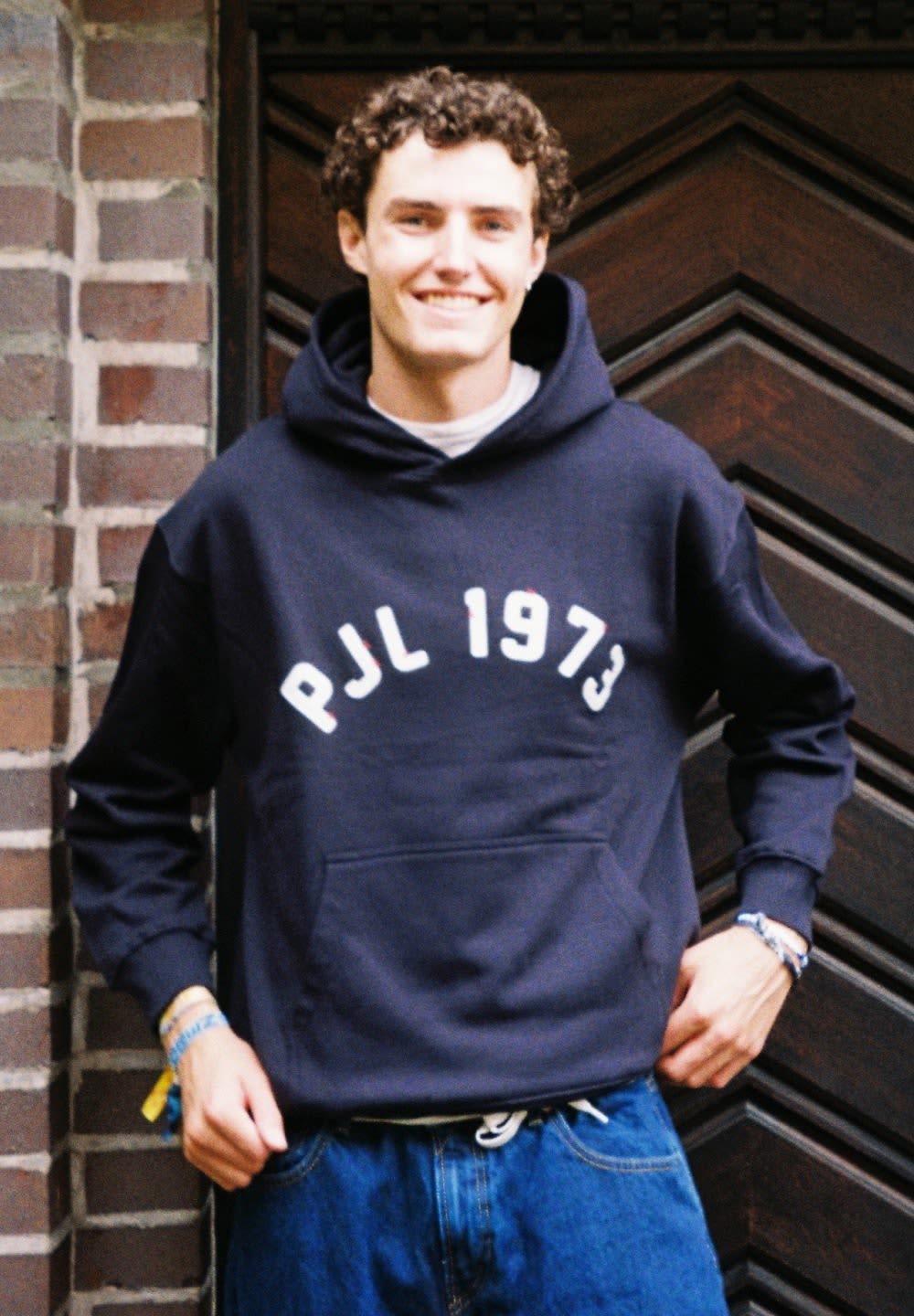 Sudadera marina con texto blanco "PJL 1973", material de algodón, bolsillo tipo canguro y capucha con cordón. Llevada con jeans de mezclilla holgados.