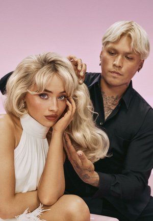 Blondt, bølget hår med pandehår på en kvinde iført en hvid, ærmeløs top. Mand i sort skjorte styler forsigtigt hendes hår mod en lyserød baggrund.