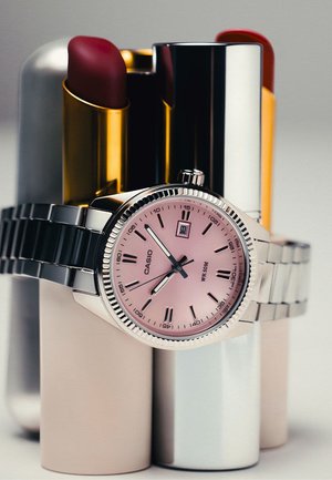 Zilveren Casio polshorloge met een roze wijzerplaat die 11:53 aangeeft, rustend op vier buisjes rode lippenstift met gouden en zilveren behuizingen.
