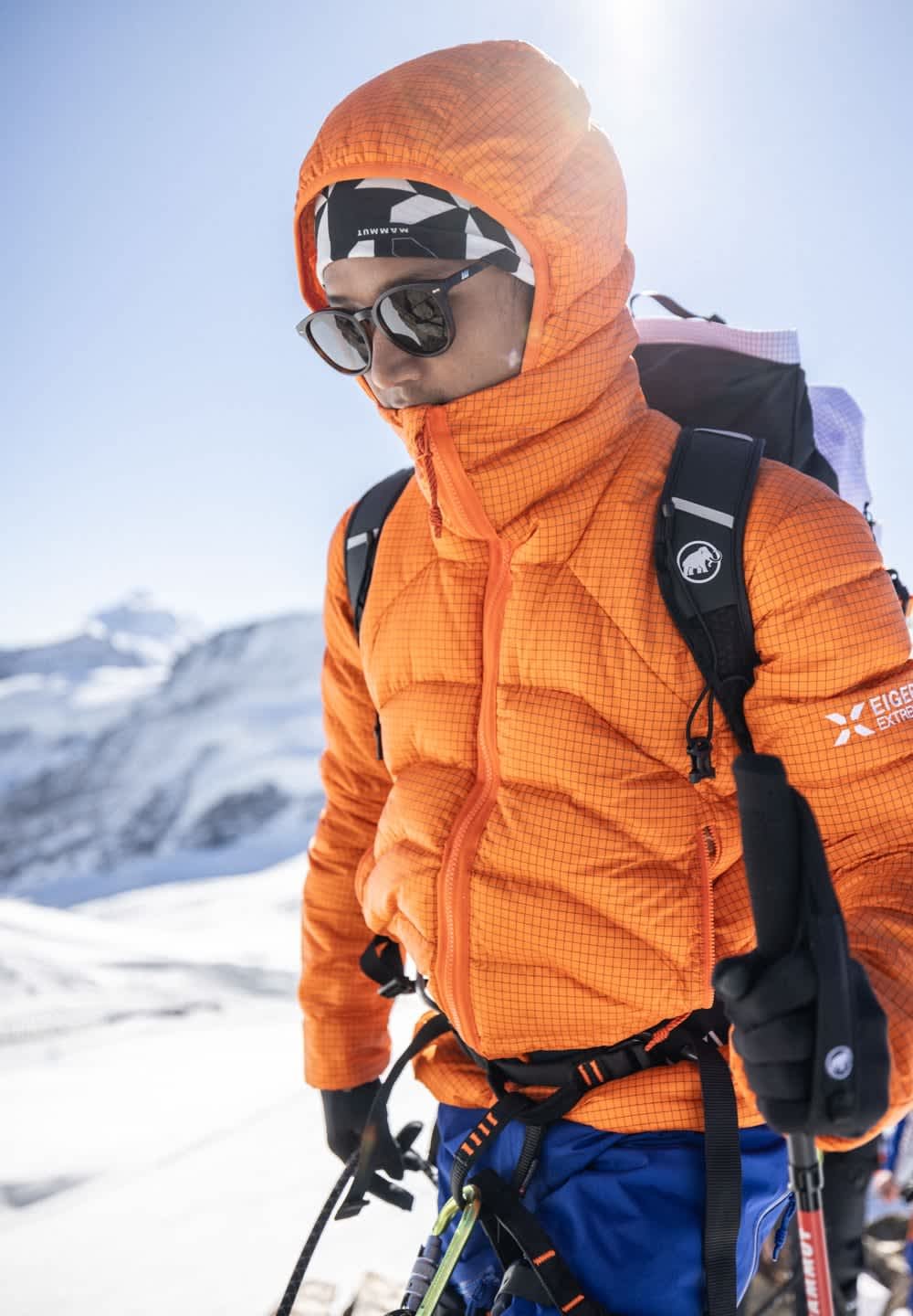 Wanderer in orange Jacke und Sonnenbrille, mit Rucksack und Trekkstöcken, steht in schneebedecktem Gebirge.