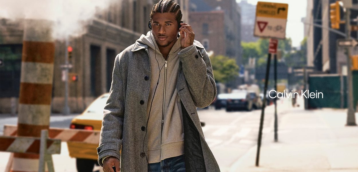 Model wearing a gray herringbone coat over a light gray hoodie, with braided hair and headphones, standing in an urban setting.