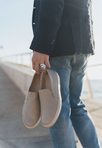 Helle beige Leder-Slipper mit strukturiertem Finish und Jute-ähnlicher Sohle, in der Hand einer Person nahe einem Küstenweg gehalten.